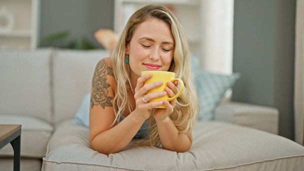 Jeune femme blonde sentant le café couché sur le canapé à la maison - Photo, image