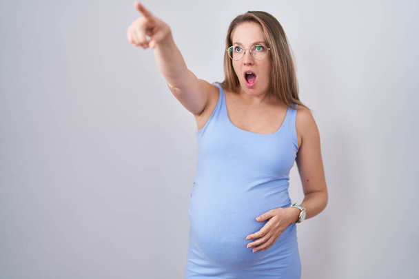 Beyaz arka planda duran hamile bir kadın parmağını öne uzatmış, şaşırmış, ağzı açık, şaşırmış, ön tarafta bir şey var.  - Fotoğraf, Görsel