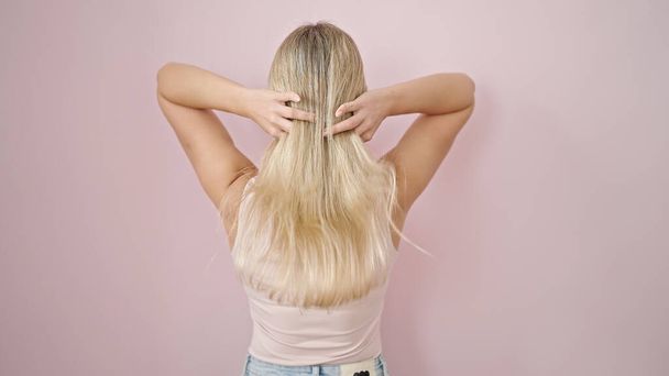 Mladá blondýna žena dotýká její vlasy pozpátku přes izolované růžové pozadí - Fotografie, Obrázek