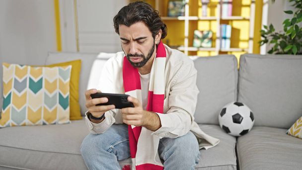 Jonge Spaanse man kijkt naar voetbalwedstrijd zittend op de bank thuis - Foto, afbeelding