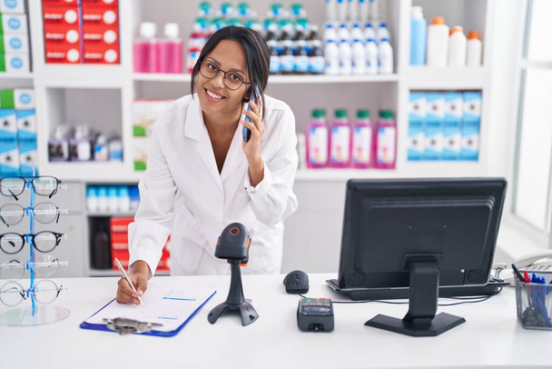 Jovem mulher hispânica farmacêutica falando no smartphone escrevendo no documento na farmácia - Foto, Imagem