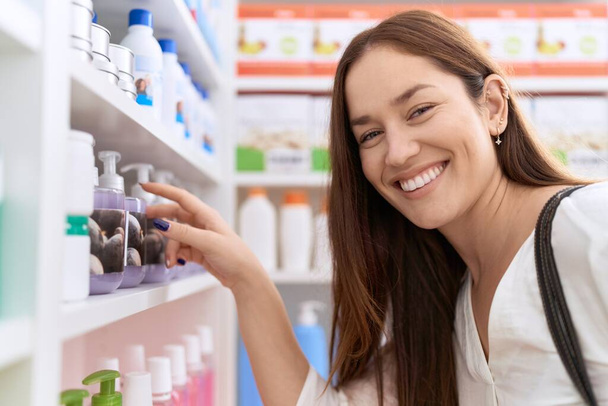 Joven hermosa mujer cliente sonriendo confiado sosteniendo el producto en estanterías en la farmacia - Foto, Imagen