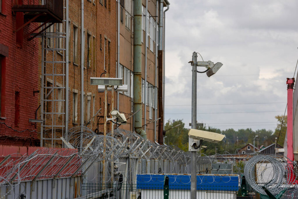 Stacheldraht und Überwachungskameras über Zaun in der Stadt nahe roter Backsteinmauer an Sommertagen, Teleaufnahme mit selektivem Fokus - Foto, Bild