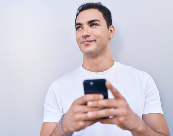 Νεαρός Ισπανόφωνος άντρας χαμογελά με αυτοπεποίθηση χρησιμοποιώντας smartphone πάνω από απομονωμένο λευκό φόντο - Φωτογραφία, εικόνα