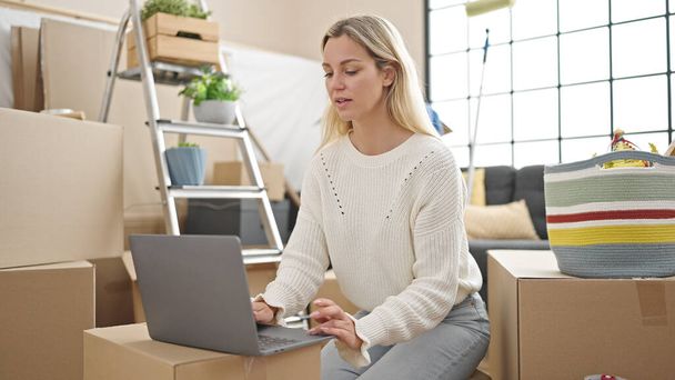 Nuori blondi nainen käyttää kannettavaa tietokonetta istuu lattialla uudessa kodissa - Valokuva, kuva