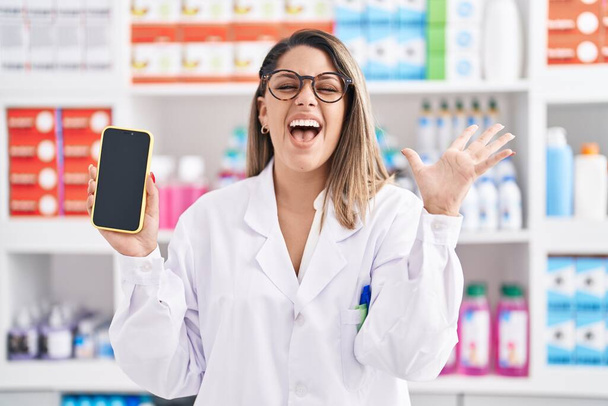 Femme blonde travaillant à la pharmacie drugstore montrant écran smartphone célébrant la victoire avec sourire heureux et expression gagnante avec les mains levées  - Photo, image