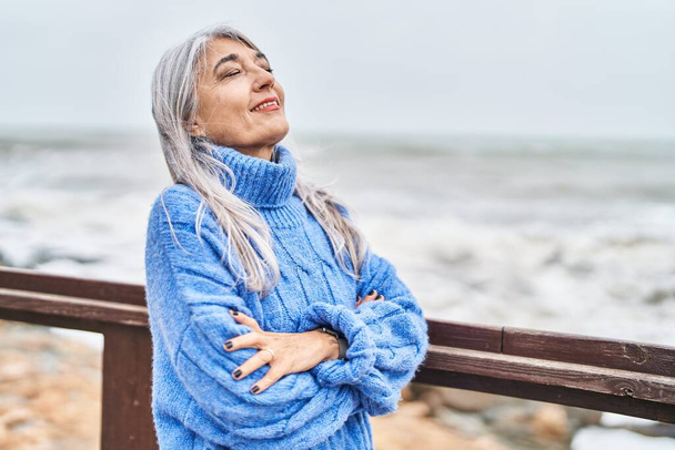 Седовласая женщина средних лет улыбается уверенно стоя со скрещенными руками на берегу моря - Фото, изображение
