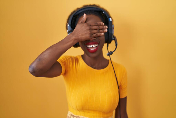 Африканська жінка з кучерявим волоссям, що стоїть над жовтим тлом, одягнена в навушники, посміхається і сміється рукою на обличчі, прикриваючи очі сюрпризом. сліпа концепція.  - Фото, зображення