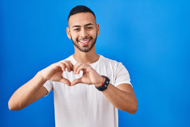 Jeune homme hispanique debout sur fond bleu souriant dans l'amour faisant forme de symbole de coeur avec les mains. concept romantique.  - Photo, image
