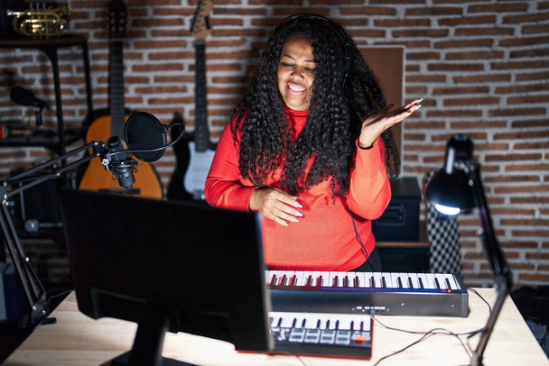 Mujer hispana de tamaño grande tocando el piano en el estudio de música sonriendo alegre presentando y señalando con la palma de la mano mirando a la cámara.  - Foto, imagen