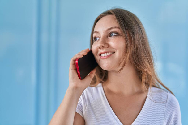 Молода блондинка посміхається впевнено розмовляючи на смартфоні над ізольованим синім фоном стіни
 - Фото, зображення