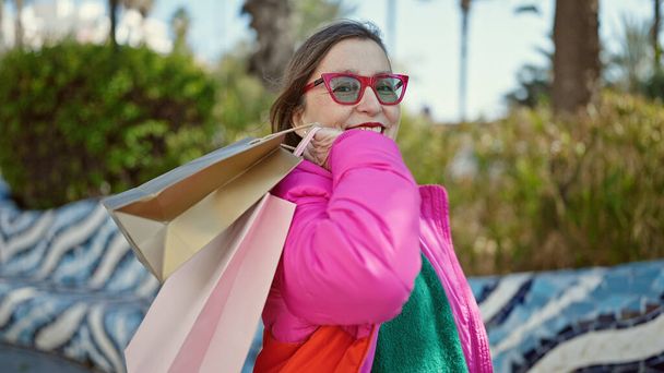 Ώριμη ισπανόφωνη γυναίκα με γκρίζα μαλλιά χαμογελώντας πηγαίνει για ψώνια κρατώντας τσάντες στο πάρκο - Φωτογραφία, εικόνα