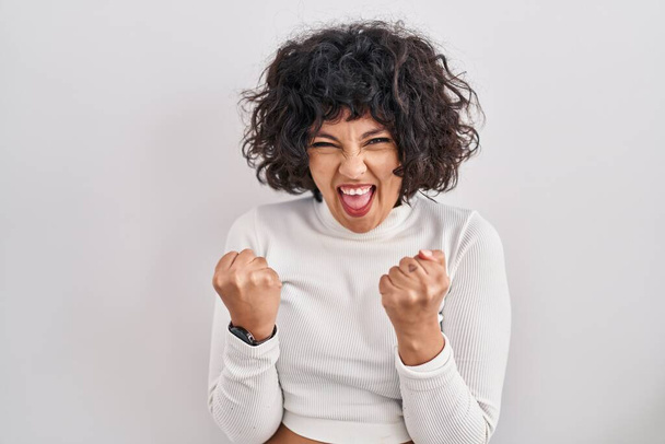Hispanic woman with curly hair standing over isolated background very happy and excited doing winner gesture with arms raised, smiling and screaming for success. celebration concept.  - Photo, Image