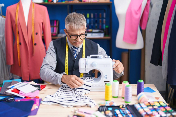 Middle age grey-haired man tailor using sewing machine at clothing factory - 写真・画像