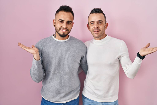 Гомосексуальная пара, стоящая на розовом фоне, весело улыбаясь и показывая ладонью на камеру.  - Фото, изображение