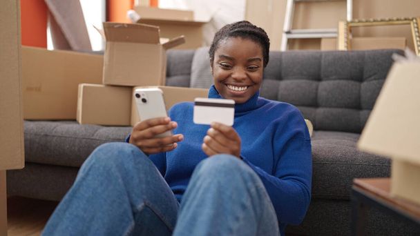 African american woman shopping with smartphone and credit card sitting on floor at new home - Foto, Bild