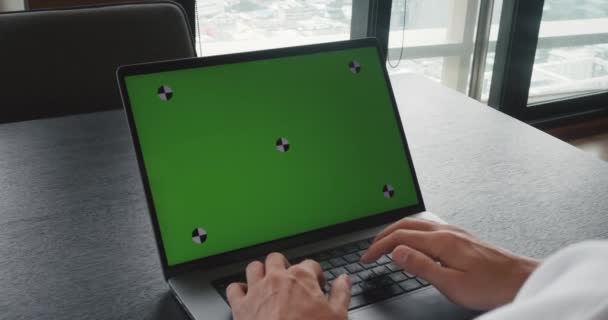 Close-up zakenman werkt op kantoor op laptop op bureau, kijkt naar groen scherm, over de schouder schot. Kantoorpersoon met laptop computer met laptop groen scherm, typt tekst op het toetsenbord. - Video