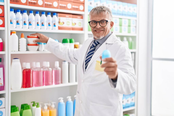 Μεσήλικας γκριζομάλλης φαρμακοποιός κρατώντας φιάλη αποσμητικού στο εργαστήριο - Φωτογραφία, εικόνα
