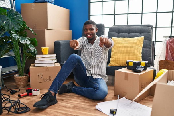 Afroamerikanischer Mann sitzt auf dem Boden im neuen Zuhause und zeigt mit den Fingern auf dich und die Kamera, lächelt positiv und fröhlich  - Foto, Bild