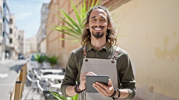 Giovane uomo ispanico cameriere sorridente fiducioso utilizzando touchpad sulla terrazza della caffetteria - Foto, immagini