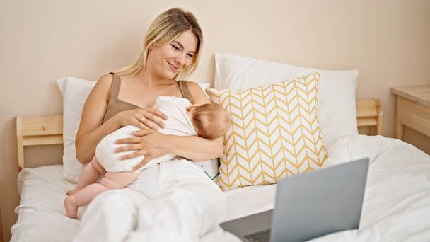 Madre e figlia sedute sul letto che allattano bambino che guarda film sul computer portatile in camera da letto - Foto, immagini