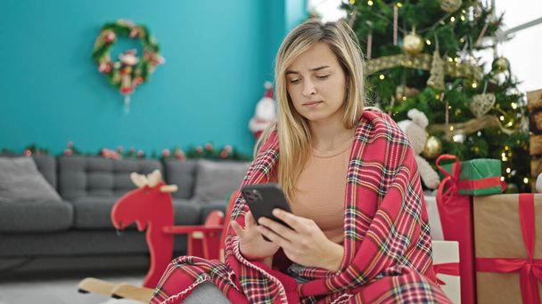 Νεαρή ξανθιά γυναίκα γιορτάζει τα Χριστούγεννα χρησιμοποιώντας smartphone αναζητούν αναστατωμένος στο σπίτι - Φωτογραφία, εικόνα