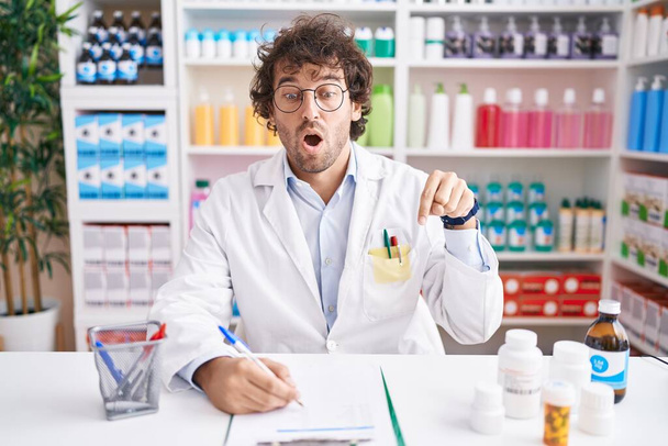 Latino nuori mies työskentelee apteekissa apteekki apteekki osoittaa alas sormet näyttää mainosta, yllättynyt kasvot ja suu auki  - Valokuva, kuva