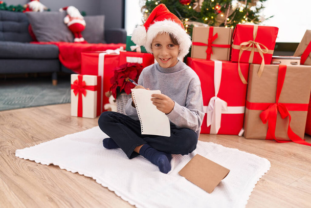 Adorable hispanic boy writing santa claus letter sitting by christmas tree at home - Foto, Bild