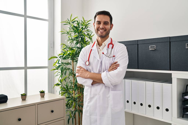 Young hispanic man wearing doctor uniform standing with arms crossed gesture at clinic - Fotografie, Obrázek