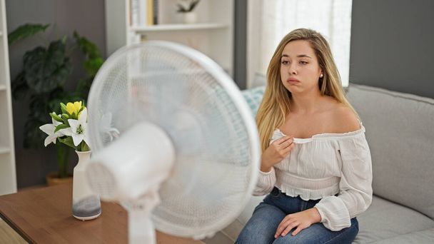 Mladá blondýna žena sedí na pohovce utrpení pro horké pomocí ventilátoru doma - Fotografie, Obrázek