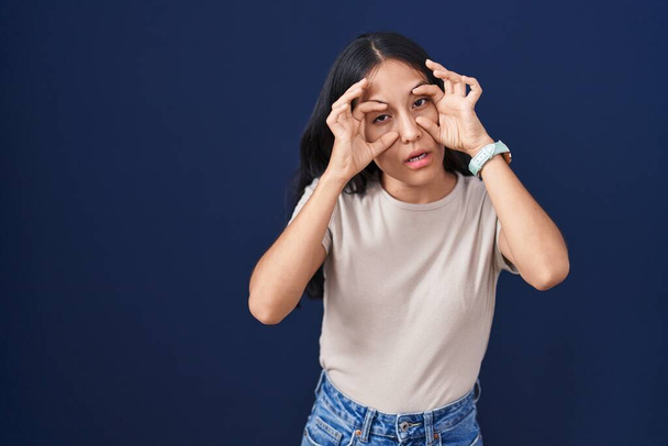 Молодая латиноамериканка, стоящая на синем фоне, пытается открыть глаза пальцами, сонная и уставшая от утренней усталости  - Фото, изображение