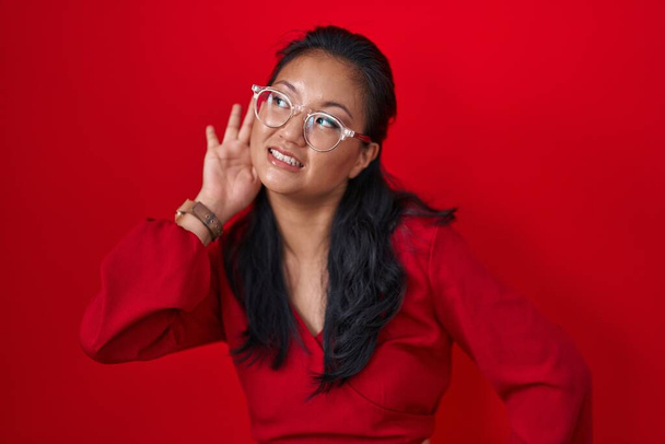 Asiatische junge Frau, die vor rotem Hintergrund steht und lächelnd mit der Hand über dem Ohr Gerüchten oder Klatsch lauscht. Konzept für Gehörlosigkeit.  - Foto, Bild