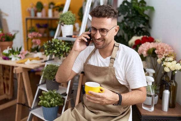 Young hispanic man florist drinking cup of coffee talking on smartphone at flower shop - Photo, Image