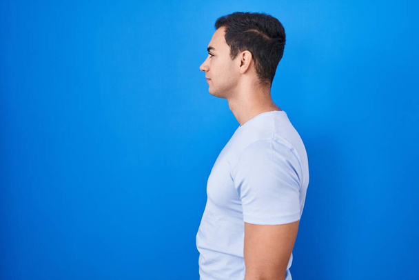 Jonge Spaanse man staande over blauwe achtergrond op zoek naar kant, ontspannen profiel poseren met een natuurlijk gezicht en zelfverzekerde glimlach.  - Foto, afbeelding