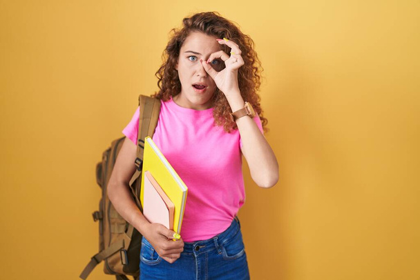 Młoda kaukaska kobieta w studenckim plecaku i trzymająca książki robiąca ok gest wstrząśnięty zaskoczoną twarzą, okiem patrzącym przez palce. niewierząca ekspresja.  - Zdjęcie, obraz