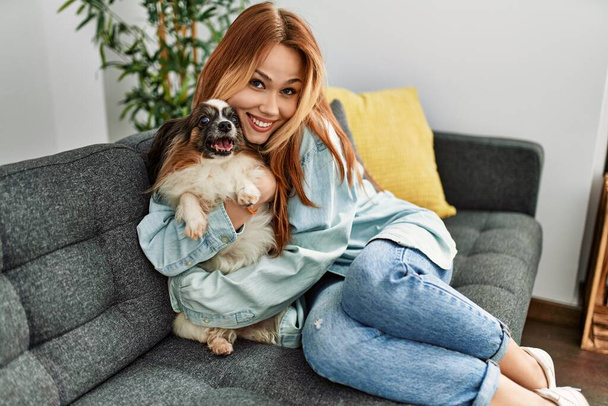 Young caucasian woman sitting on sofa hugging dog at home - Photo, Image