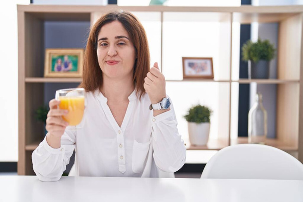 Brunette woman drinking glass of orange juice doing money gesture with hands, asking for salary payment, millionaire business  - Photo, Image