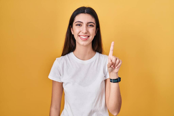 Young beautiful woman standing over yellow background showing and pointing up with finger number one while smiling confident and happy.  - Photo, Image