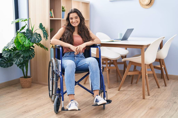 jong tiener meisje zitten op rolstoel in de woonkamer kijken positief en gelukkig staan en glimlachen met een zelfverzekerde glimlach tonen tanden  - Foto, afbeelding