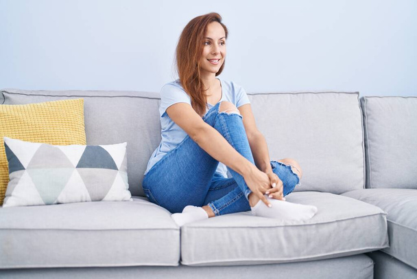 Νεαρή γυναίκα χαμογελά αυτοπεποίθηση κάθεται στον καναπέ στο σπίτι - Φωτογραφία, εικόνα
