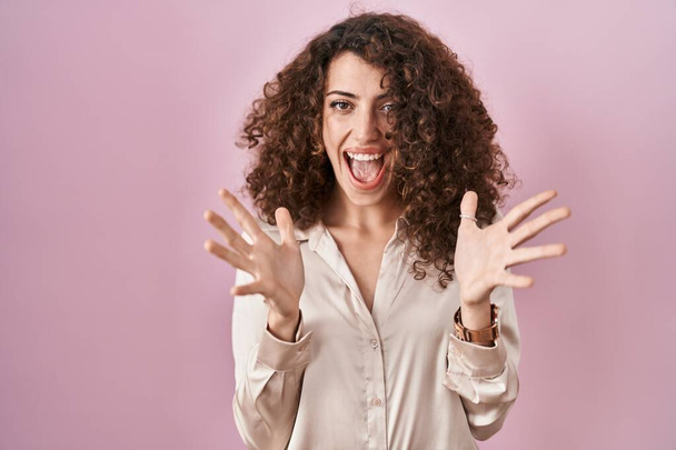 Hispanic woman with curly hair standing over pink background celebrating crazy and amazed for success with arms raised and open eyes screaming excited. winner concept  - Fotoğraf, Görsel