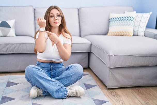 Молода біла жінка сидить на підлозі у вітальні, поспішаючи вказуючи на час, нетерплячість, дивлячись на камеру з розслабленим виразом  - Фото, зображення