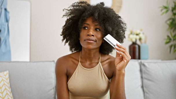 Afroamerikanerin mit Kreditkarte sitzt zu Hause auf Sofa und denkt nach - Foto, Bild