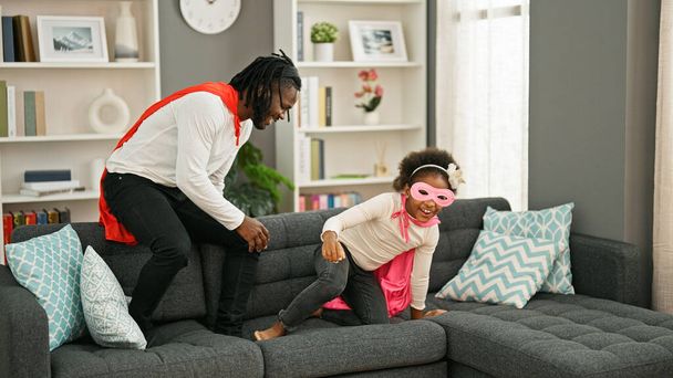 Afroamericano padre e figlia sorridente fiducioso indossare costume da supereroe giocare sul divano a casa - Foto, immagini