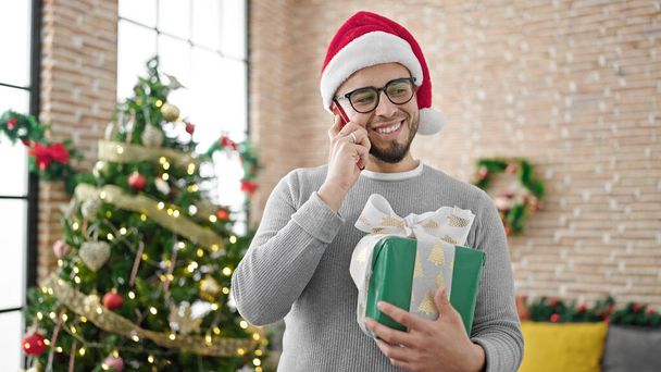 Hispanic man wearing christmas hat holding gift speaking on the phone at home - Photo, Image