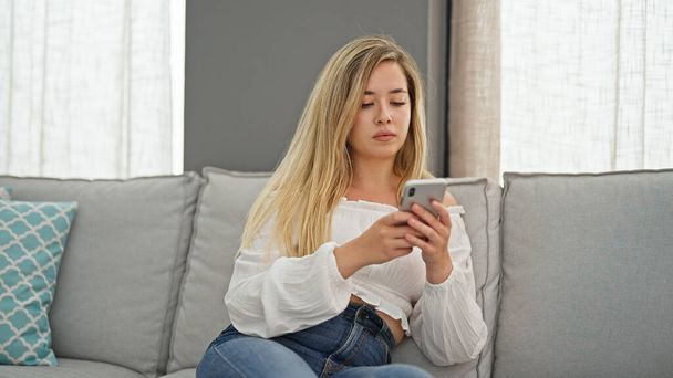 Młoda blondynka za pomocą smartfona siedzi na kanapie myśląc w domu - Zdjęcie, obraz