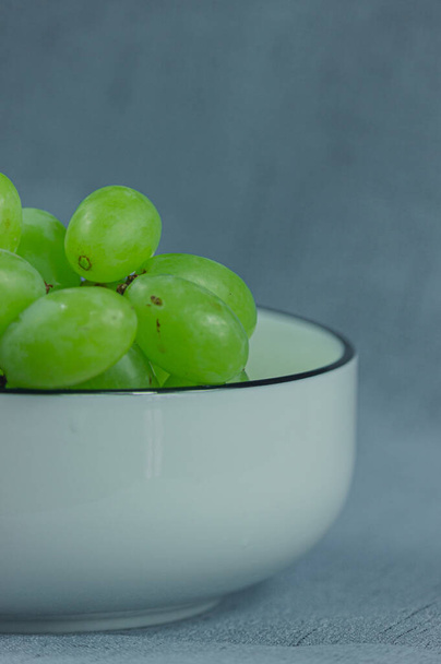Fresh green grapes in a white ceramic bowl on a solid color background. In the vertical composition is a close-up of the white ceramic bowl and part of the grapes - Photo, Image