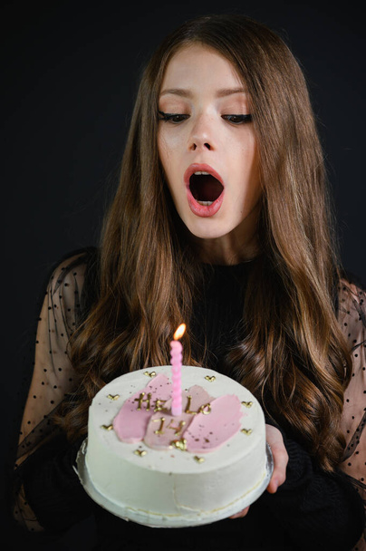 Hermosa chica adolescente feliz con vestido negro soplando una vela en un pastel de cumpleaños. Pastel con la inscripción Hello 14. - Foto, Imagen