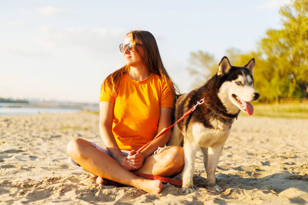Весела красива молода жінка в сонцезахисних окулярах сидить зі своєю собакою на пляжі і насолоджується заходом сонця - Фото, зображення