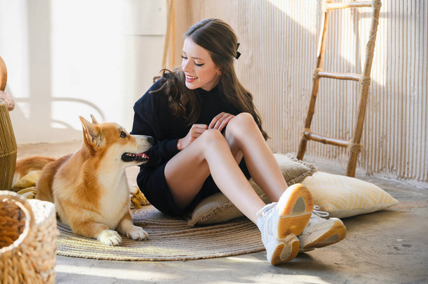 Ein Mädchen im Teenageralter sitzt mit ihrem Corgi-Hund auf dem Boden. Das Mädchen trägt ein schwarzes Sweatshirt und lange lockige Haare. - Foto, Bild